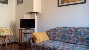 Ett vardagsrum med en mönstrad soffa, en tv på ett tv-bord, ett litet bord med ett blommigt bordsduk och ett väggmonterat skåp.