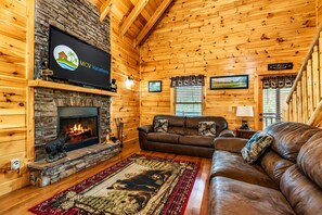 Ein gemütliches Blockhaus-Interieur mit Kamin aus Stein, einem Ledersofa, einem an der Wand montierten Fernseher und einer Holzbalkendecke.