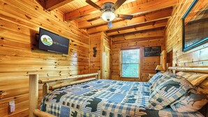 Een houten kamer met een bed, een plafondventilator, een tv aan de muur en een raam met jaloezieën.