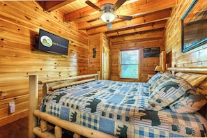 Una habitación tipo cabaña de madera con cama, ventilador de techo, televisión en la pared y ventana con persianas.
