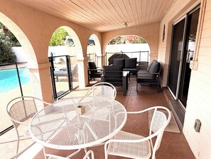 An outdoor patio with a pool, white metal chairs, a round table, and a barbecue area.