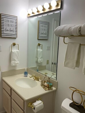 A bathroom with a vanity, a sink, a toilet, and a mirror with lights above. There are towels hanging on the wall and a gold-colored towel rack.