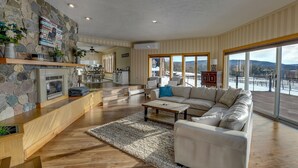 A spacious living room with a stone fireplace, a large sofa, and a view of a deck and mountains. - Nunda Ranch near Letchworth State Park (Nunda)