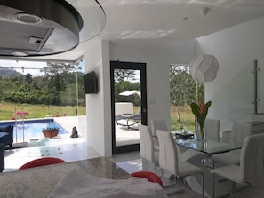 A modern kitchen with a glass dining table, white chairs, and a view of a swimming pool and garden.