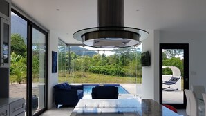 A modern kitchen with a glass-top table, a built-in stove, and a view of a swimming pool and greenery outside. - House of Views (La Fortuna)