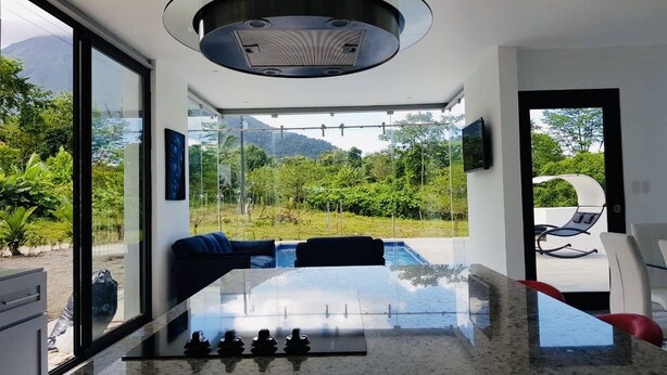 A modern kitchen with a glass-top dining table, a built-in stove, and a view of a lush garden and mountains. - House of Views (La Fortuna)