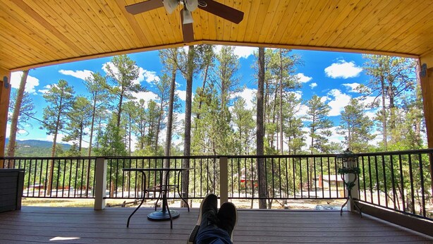 A wooden deck with a table and chairs, a railing, and a view of tall trees and a clear sky. - Cozy, Rustic Pine Ridge Cabin with beautiful views - Midtown Ruidoso (Ruidoso)