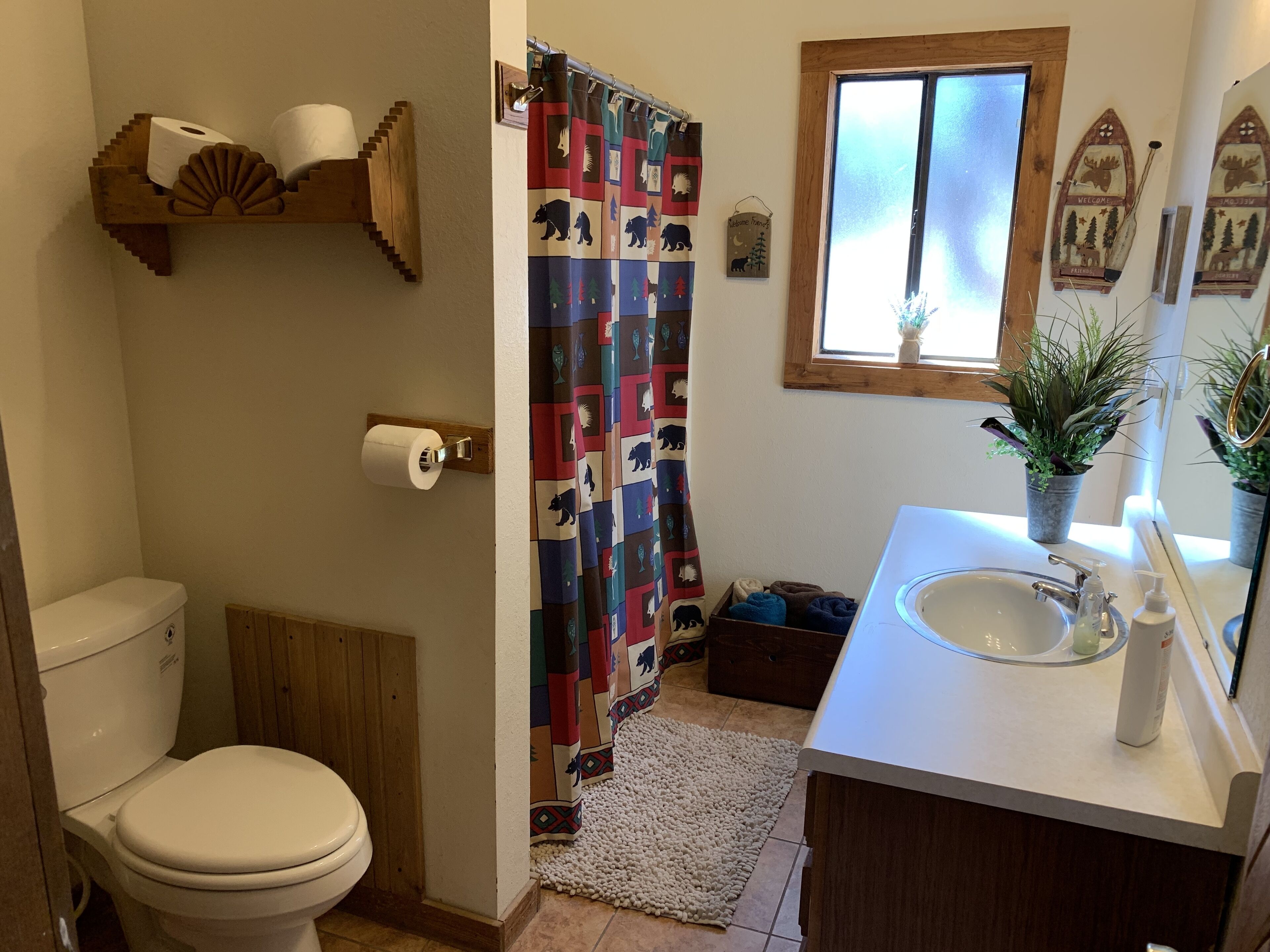 Un baño con inodoro, cortina de ducha con estampado de animales, lavabo con espejo y repisa de madera con papel higiénico.