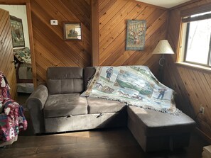 Une pièce chaleureuse avec un canapé en cuir marron, une couverture matelassée, des murs en bois, un tableau encadré et une fenêtre.