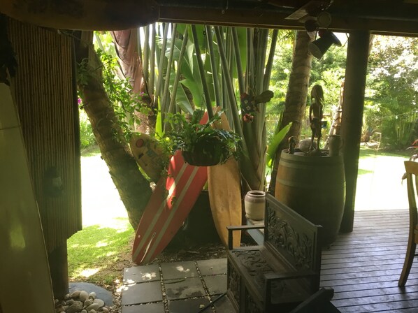 A covered outdoor area with a wooden bench, a potted plant, and a surfboard leaning against a bamboo structure.