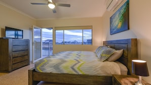 Une chambre à coucher avec un lit, une télévision, un ventilateur de plafond, une fenêtre donnant sur un beau paysage et un tableau accroché au mur.