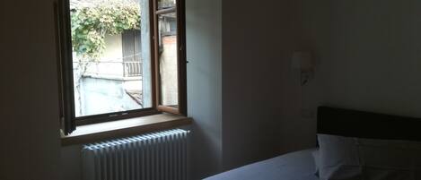 Un dormitorio con una cama individual, una ventana con contraventanas abiertas y un radiador debajo de la ventana.