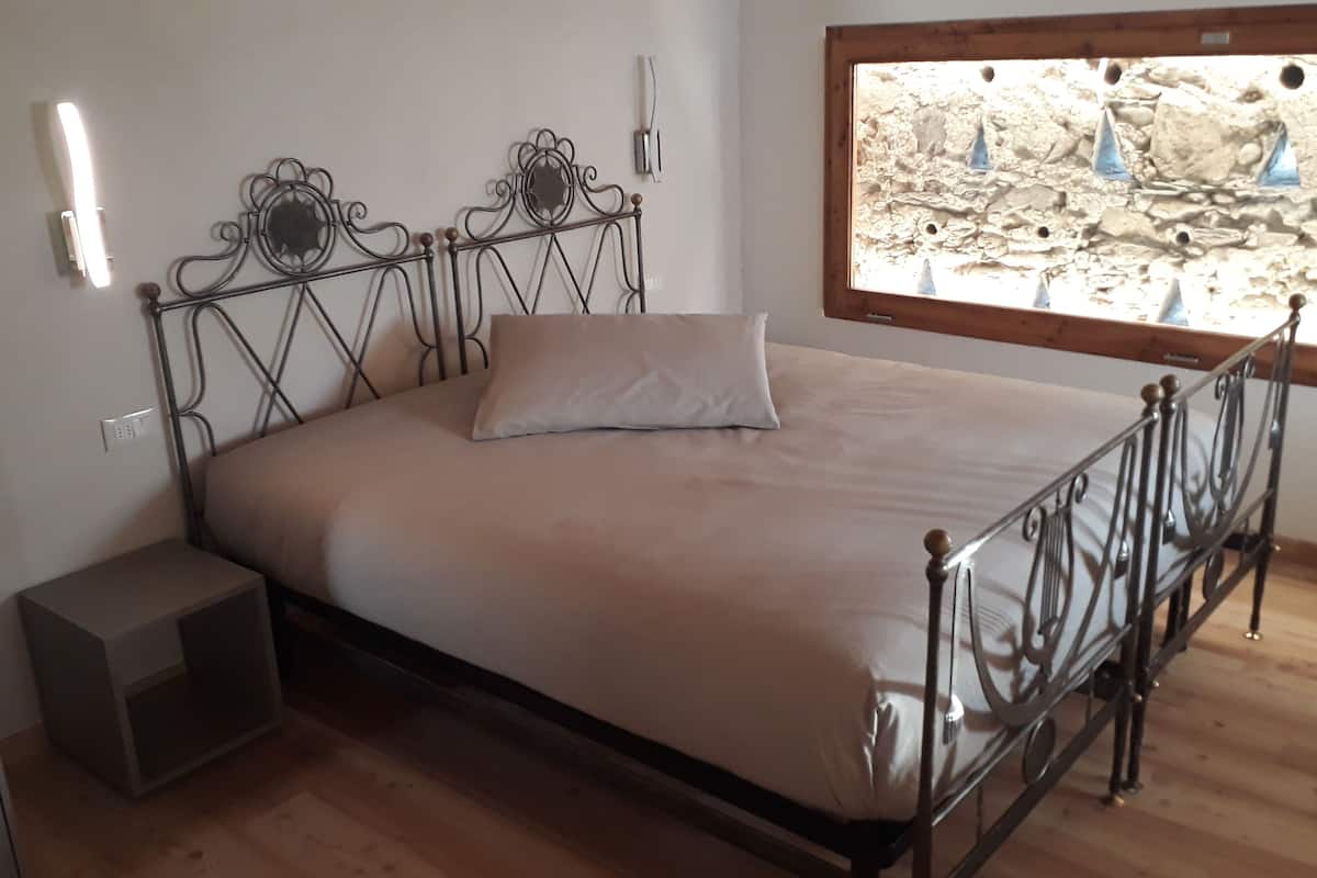 Una cama individual con estructura metálica, una mesita de noche, una lámpara fijada a la pared y una ventana con vista a un paisaje rocoso.