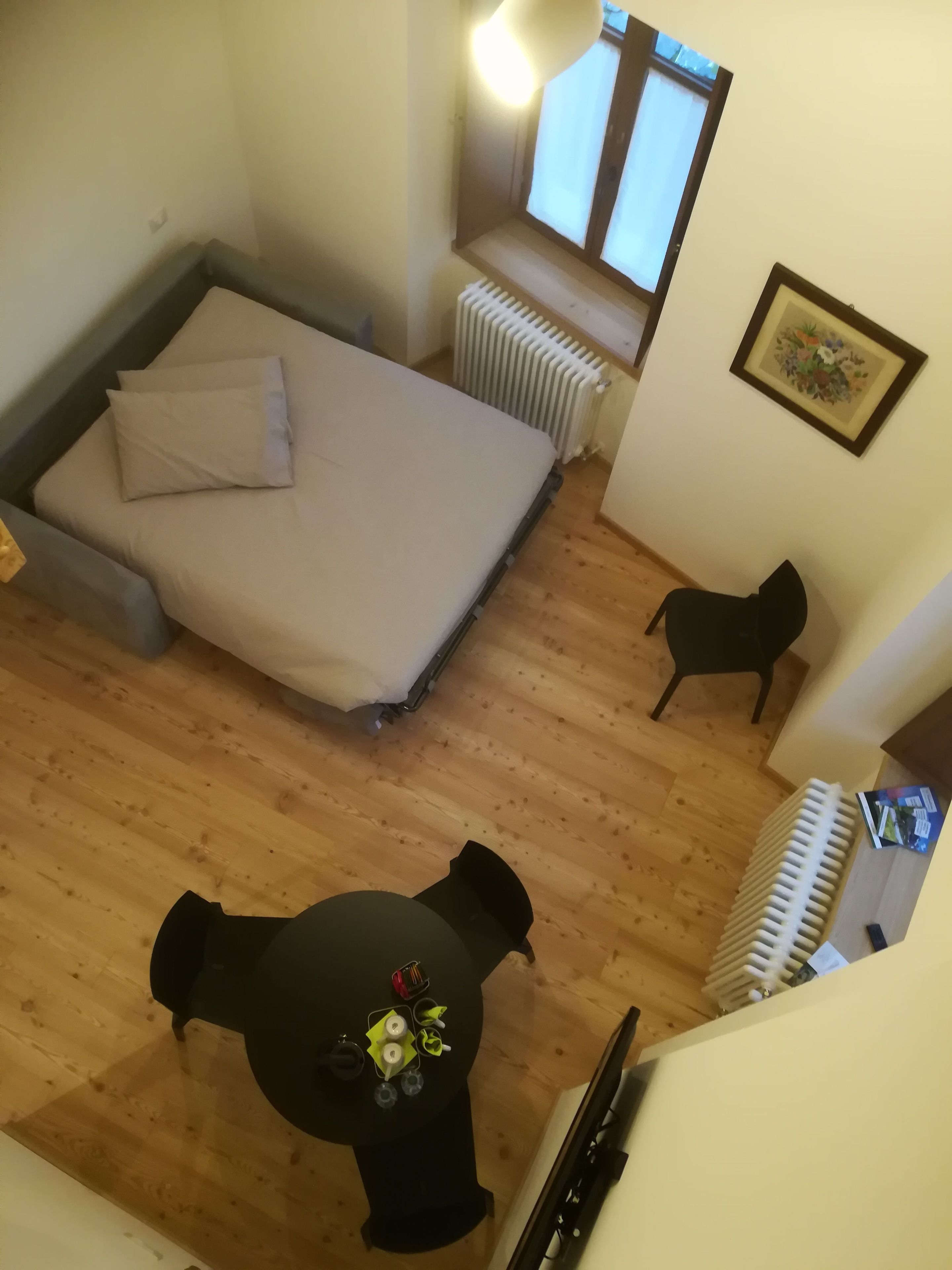 Un dormitorio con una cama, un radiador, una ventana, un cuadro enmarcado y una mesa con una silla.