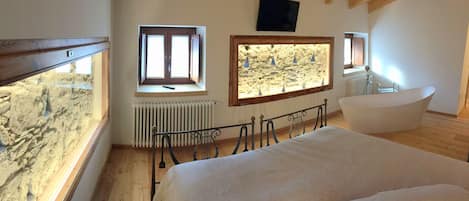 A bedroom with a bed, a window with a view, a stone wall, and a bathtub.