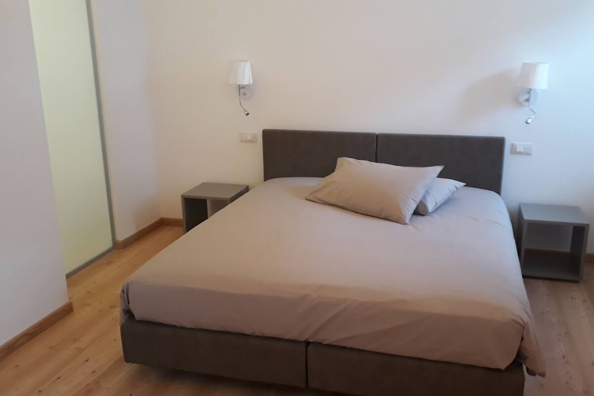 Una cama individual con cabecera gris, dos mesitas de noche y dos lámparas de pared.