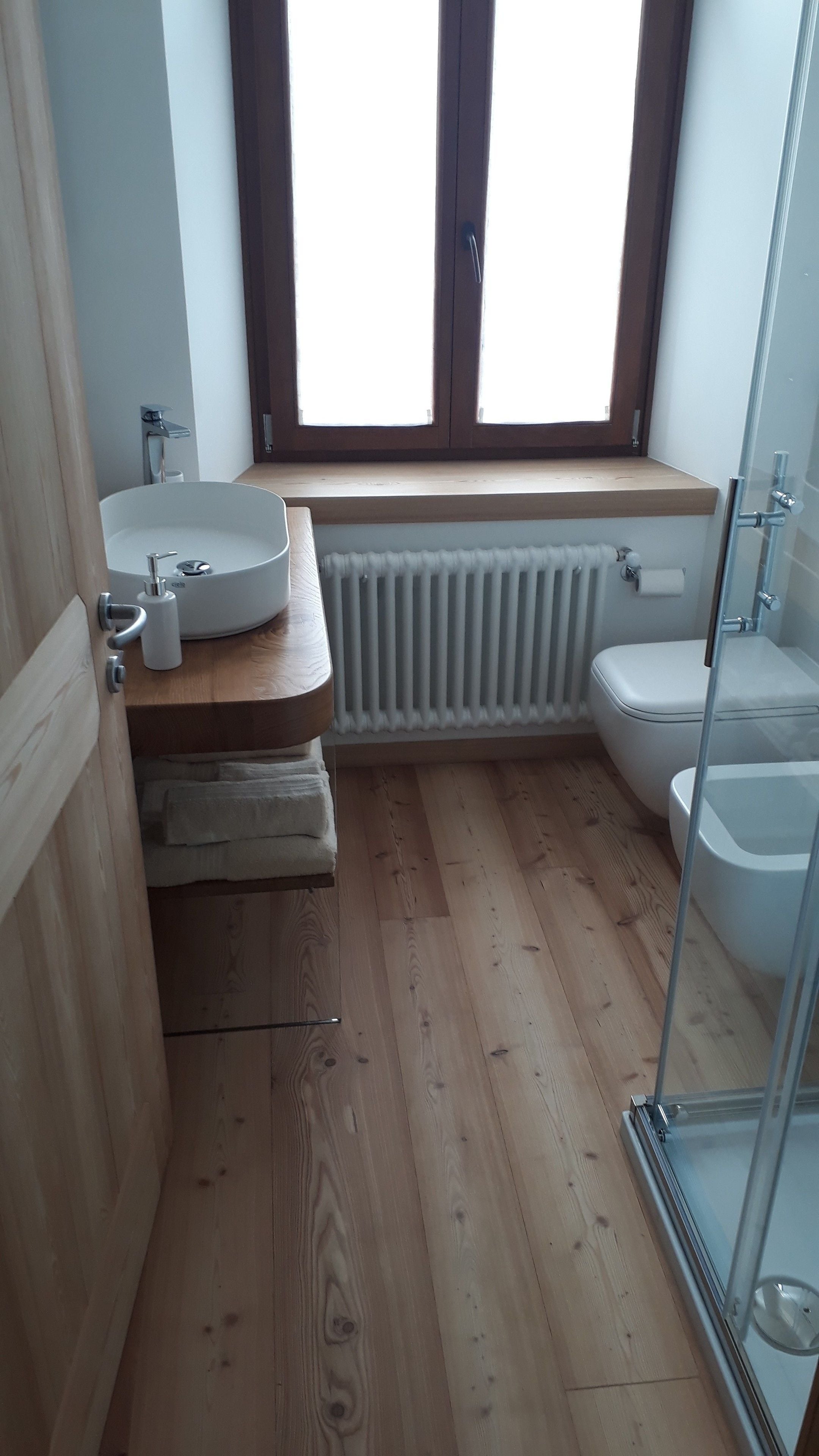 A modern bathroom with a wooden floor, a round sink, a toilet, and a glass shower enclosure.