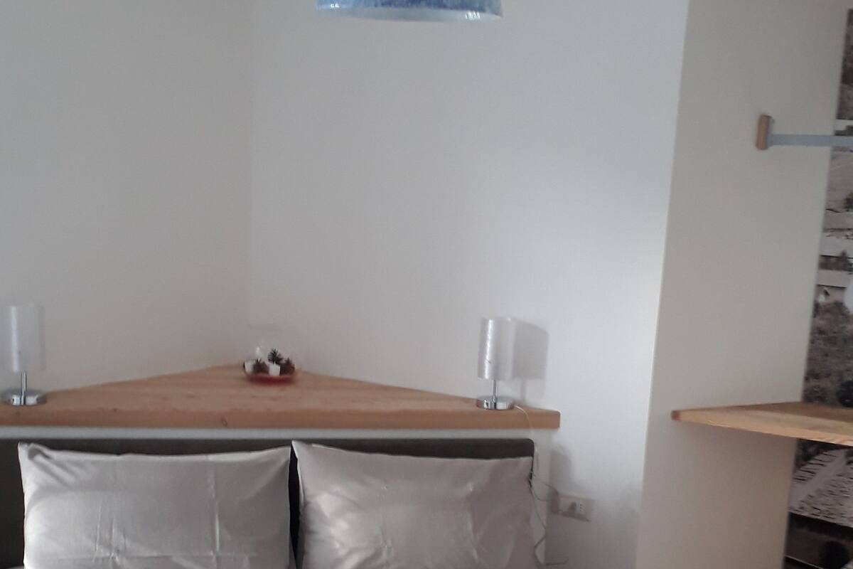 Un dormitorio con una cama, almohadas, un cabecero de madera, una lámpara colgante azul y un pequeño estante con una lámpara.