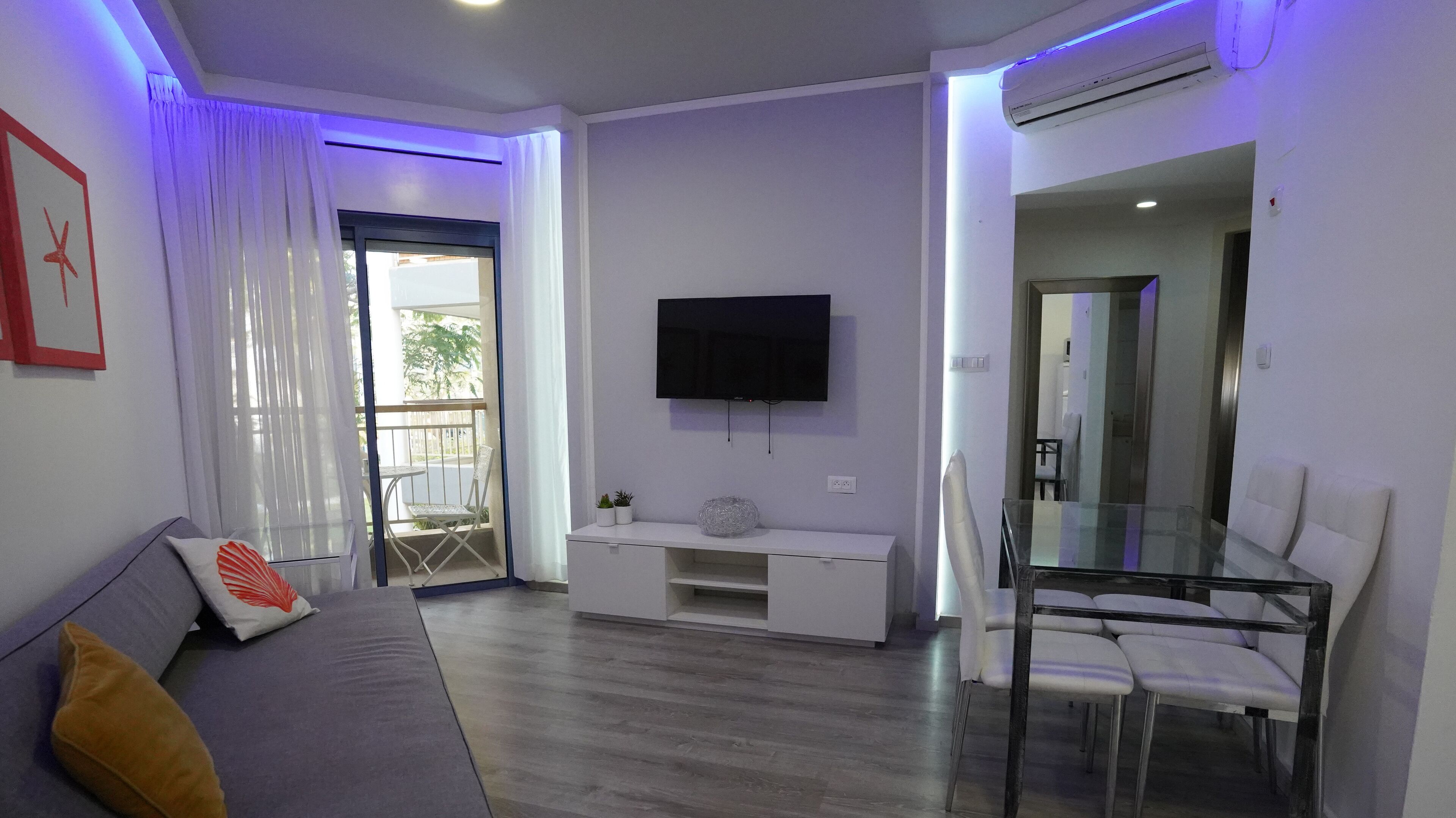 Un salon moderne avec un canapé, un téléviseur fixé au mur, une table à manger en verre et un balcon avec une chaise.