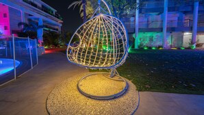 A hanging chair with a wireframe design, placed on a circular mosaic base, in an outdoor setting with a building in the background.