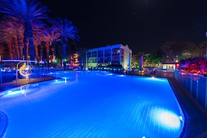 Area piscina con edificio in vetro, palme e luci colorate.