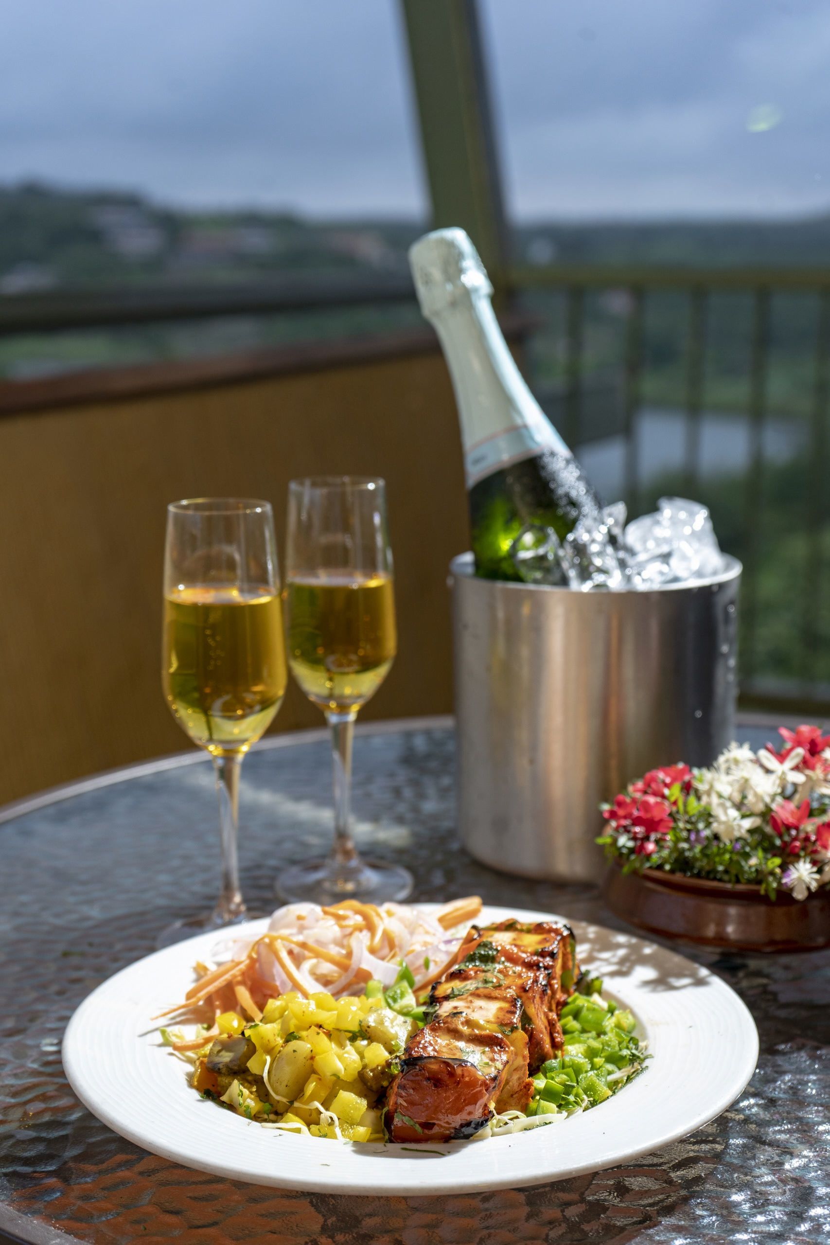 A table with a plate of food, two glasses of wine, a bottle of champagne, and a flower arrangement.