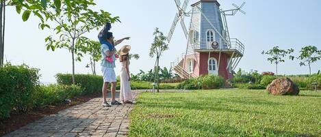 A pathway leading to a windmill with a red brick base and white upper structure, surrounded by greenery and a clear sky.