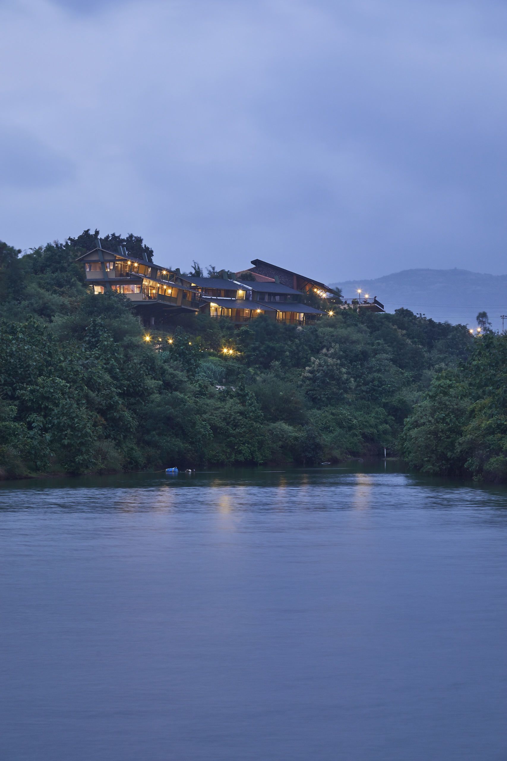 A large, multi-story building with multiple illuminated windows, situated on a hillside overlooking a body of water.
