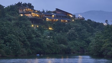 A large, multi-story building with multiple illuminated windows, situated on a hillside overlooking a body of water.