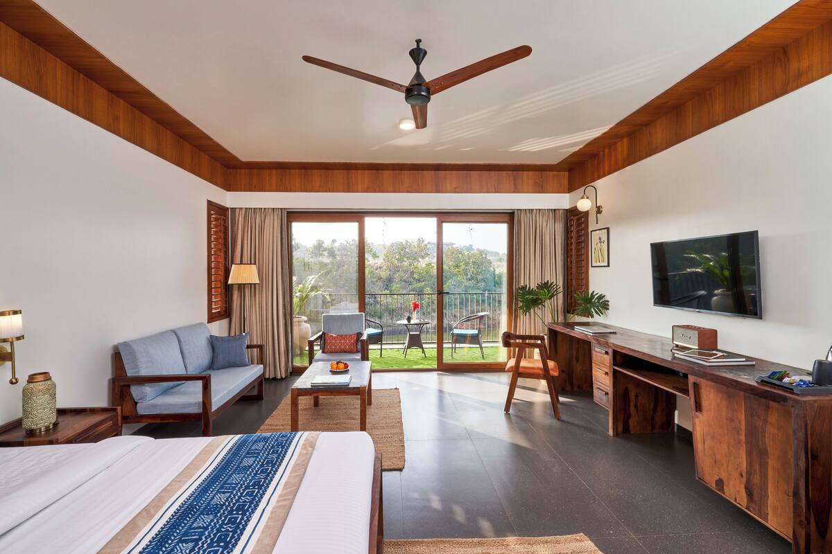 A hotel room with a bed, a desk, a sofa, and a balcony with a view of greenery.