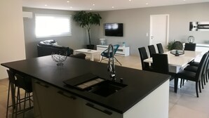 A modern kitchen with a central island, dining area with chairs, a TV mounted on the wall, and a potted plant.