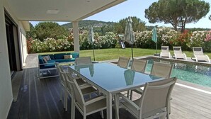 A poolside area with a glass table and chairs, a blue sofa, and a view of a garden with flowers and trees.
