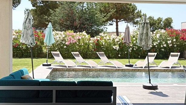 A poolside area with lounge chairs and umbrellas, a small pool, and a garden with flowers and trees.