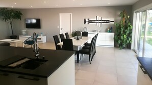 A modern kitchen with a central island, dining area with chairs, a TV mounted on the wall, and a large window providing natural light.
