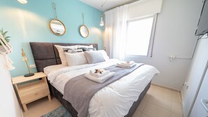 Una cama bien hecha con sábanas blancas, un plaid gris y una toalla doblada al lado. Hay una mesita de noche con una lámpara, un espejo y una pequeña planta. La habitación tiene una pared azul claro, una ventana con cortinas blancas y un suelo de baldosas.