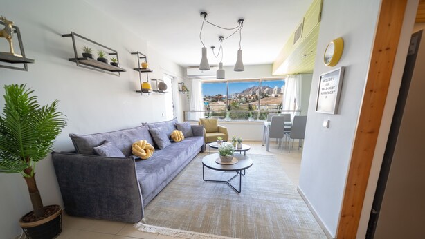 Una sala moderna con un sofá gris, una mesa de centro redonda y una planta en maceta. Hay estanterías con objetos decorativos y vistas a un paisaje urbano a través de amplias ventanas.