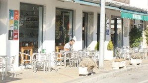 Un café al aire libre con sillas y mesas de metal, una persona sentada en una mesa y un letrero de hostal al fondo.