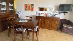 A dining area with a wooden table and chairs, a cabinet with glass doors, a wall-mounted TV, and a small desk with books and a plant. - Apartment/ flat -Cala Gonone  - Casa dolce vista - Via delle Ginestre 20 (Cala Gonone)