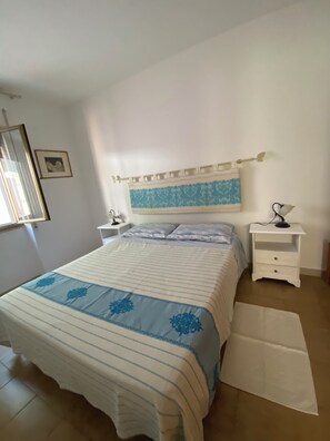 Ein Schlafzimmer mit einem Bett, einem Nachttisch, einer Lampe, einem Bild an der Wand und einem Fenster.