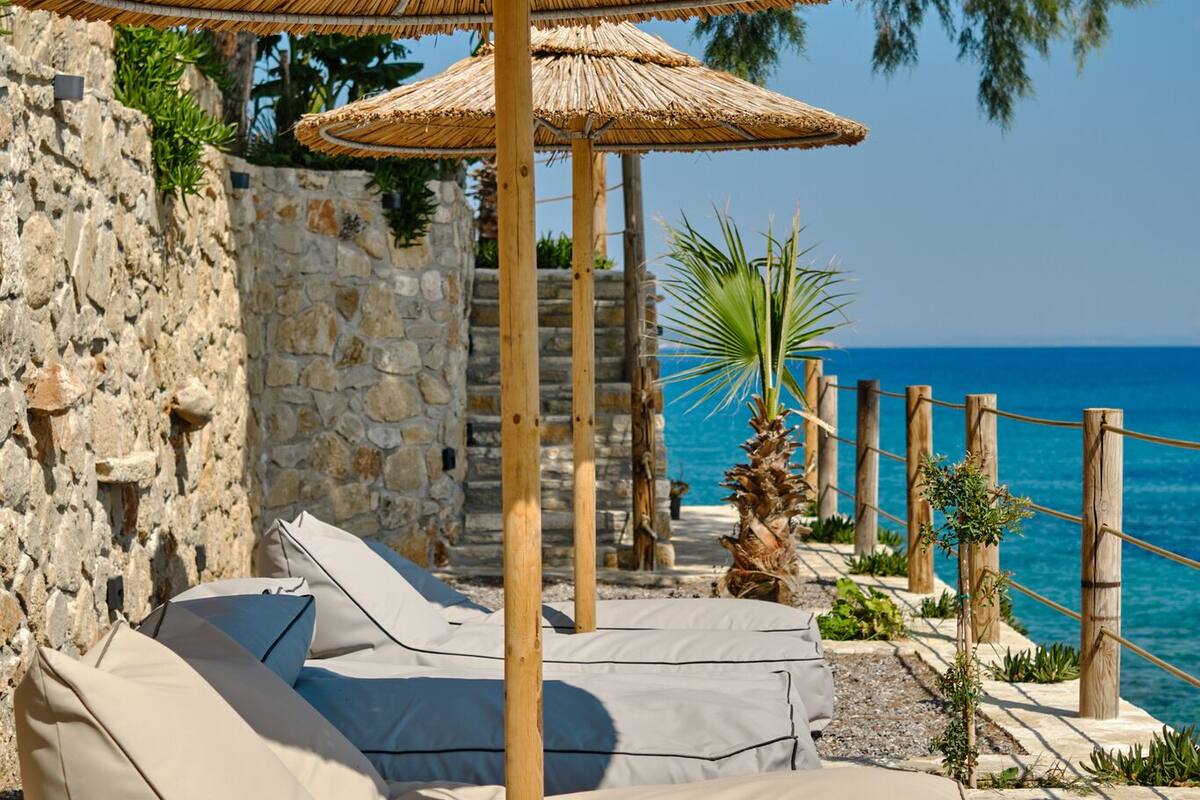 Ein Strand-Loungesbereich mit Reetdachschirmen, Steinmauern und Blick auf das Meer.