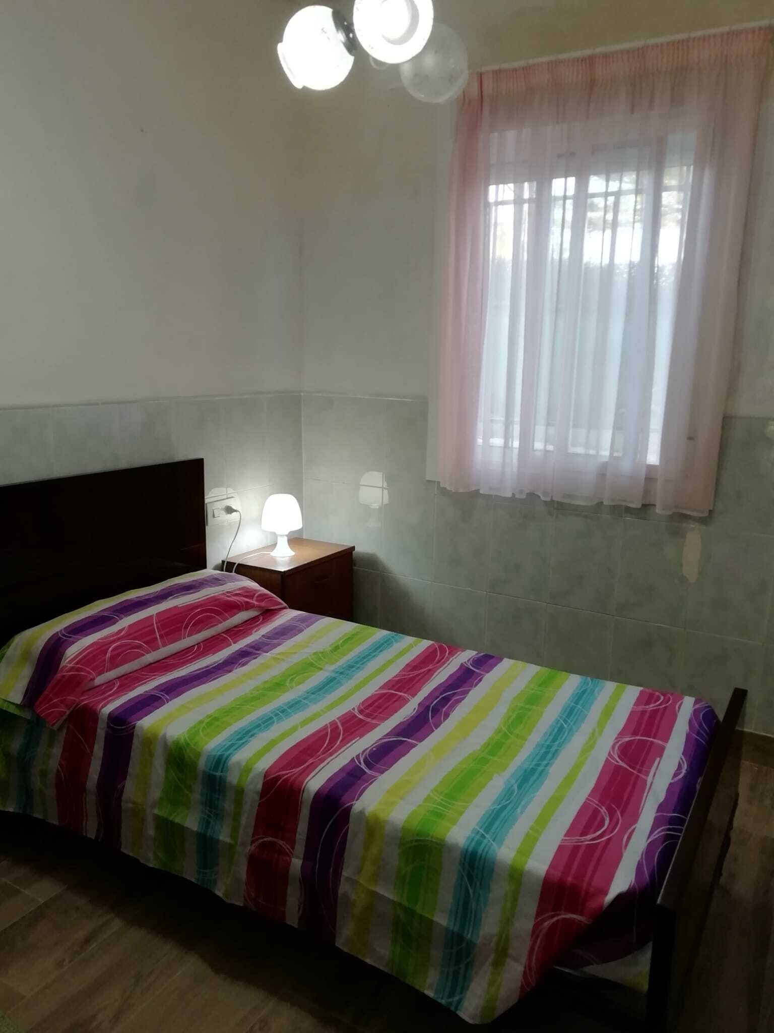 Une petite chambre avec un lit simple, une couette à rayures colorées, une table de chevet avec une lampe et une fenêtre ornée de rideaux légers.