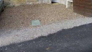 A rustic stone building with a small window and door, surrounded by gravel and greenery.