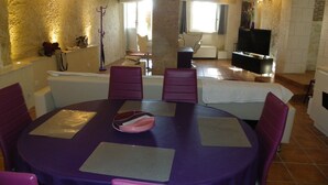 A dining area with a round table and purple chairs, a sofa, and a television in a stone-walled room.