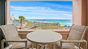 A balcony with wicker chairs and a small table, overlooking a beach with umbrellas and a flag. - Beach Front at Jetty East! One FREE Beach Chair Setup Each Day of Your Stay! (Destin)