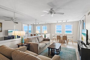 Une chambre d’hôtel comprenant un canapé, une table basse, une télévision, un coin repas avec des chaises et une vue sur la plage par les fenêtres.
