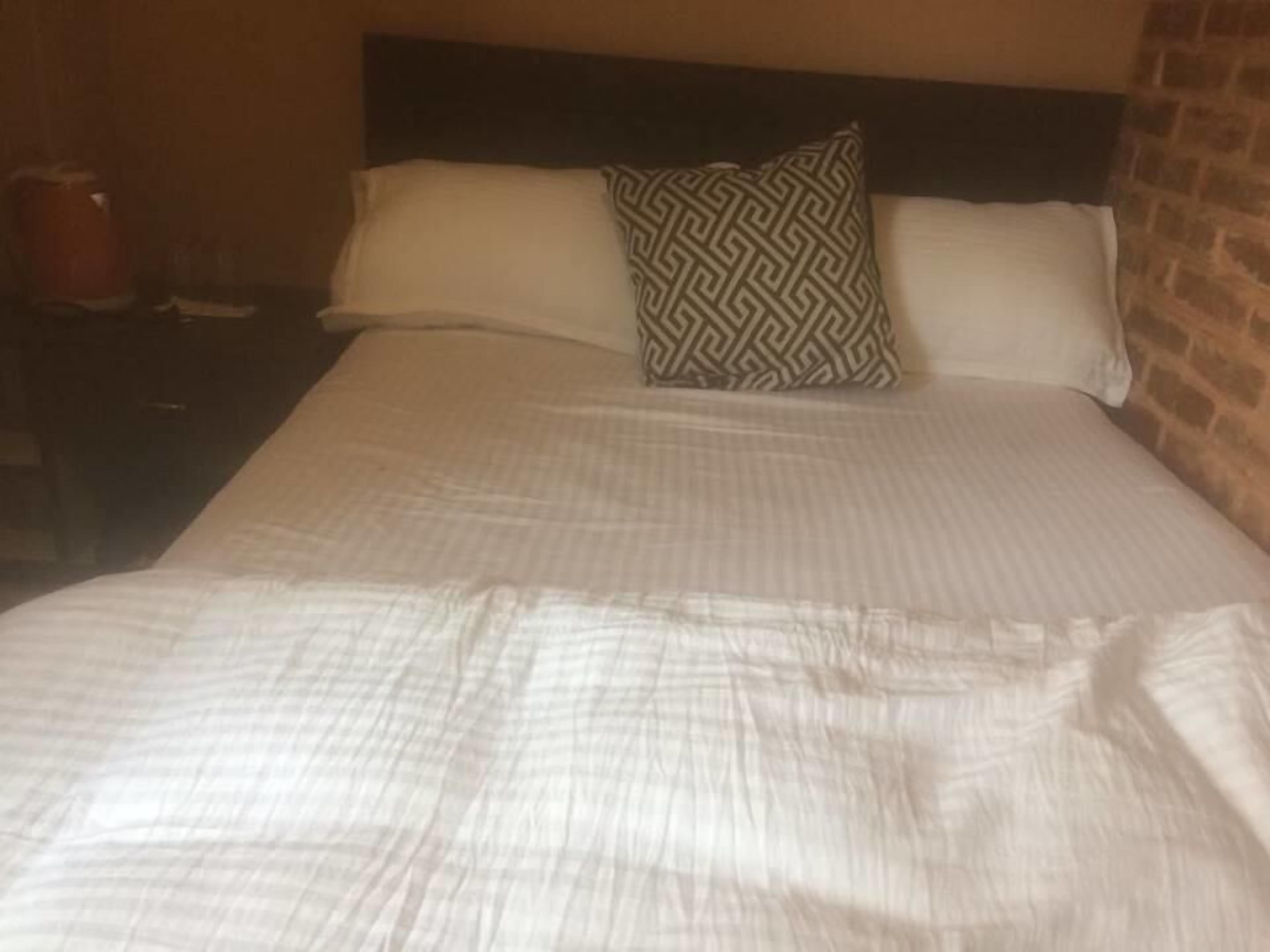 A neatly made bed with a patterned pillow and a plain pillow, a wooden nightstand with a red object on it, and a brick wall.