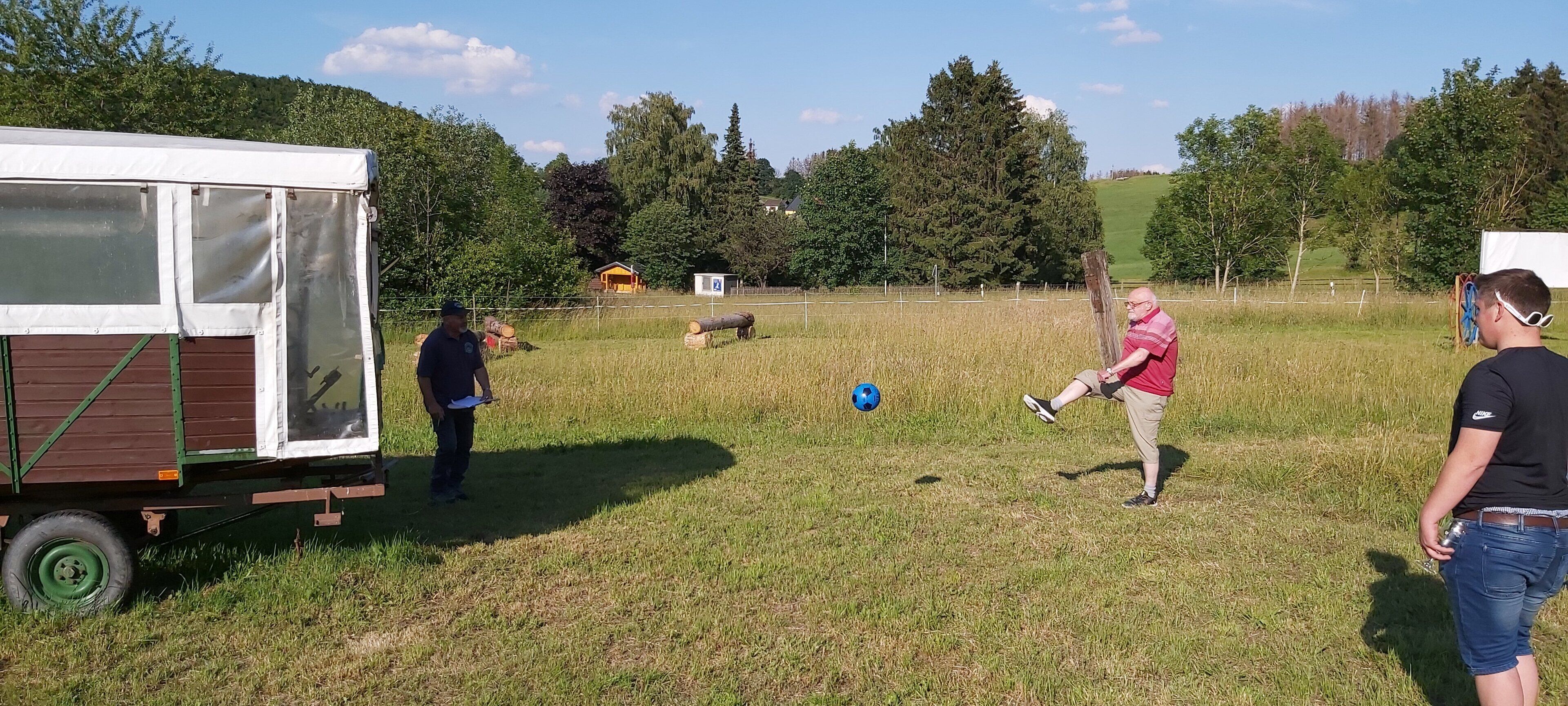 A grassy field with a trailer, a person kicking a soccer ball, and another person observing.