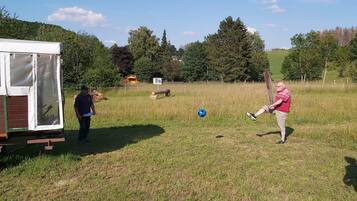 A grassy field with a trailer, a person kicking a soccer ball, and another person observing.