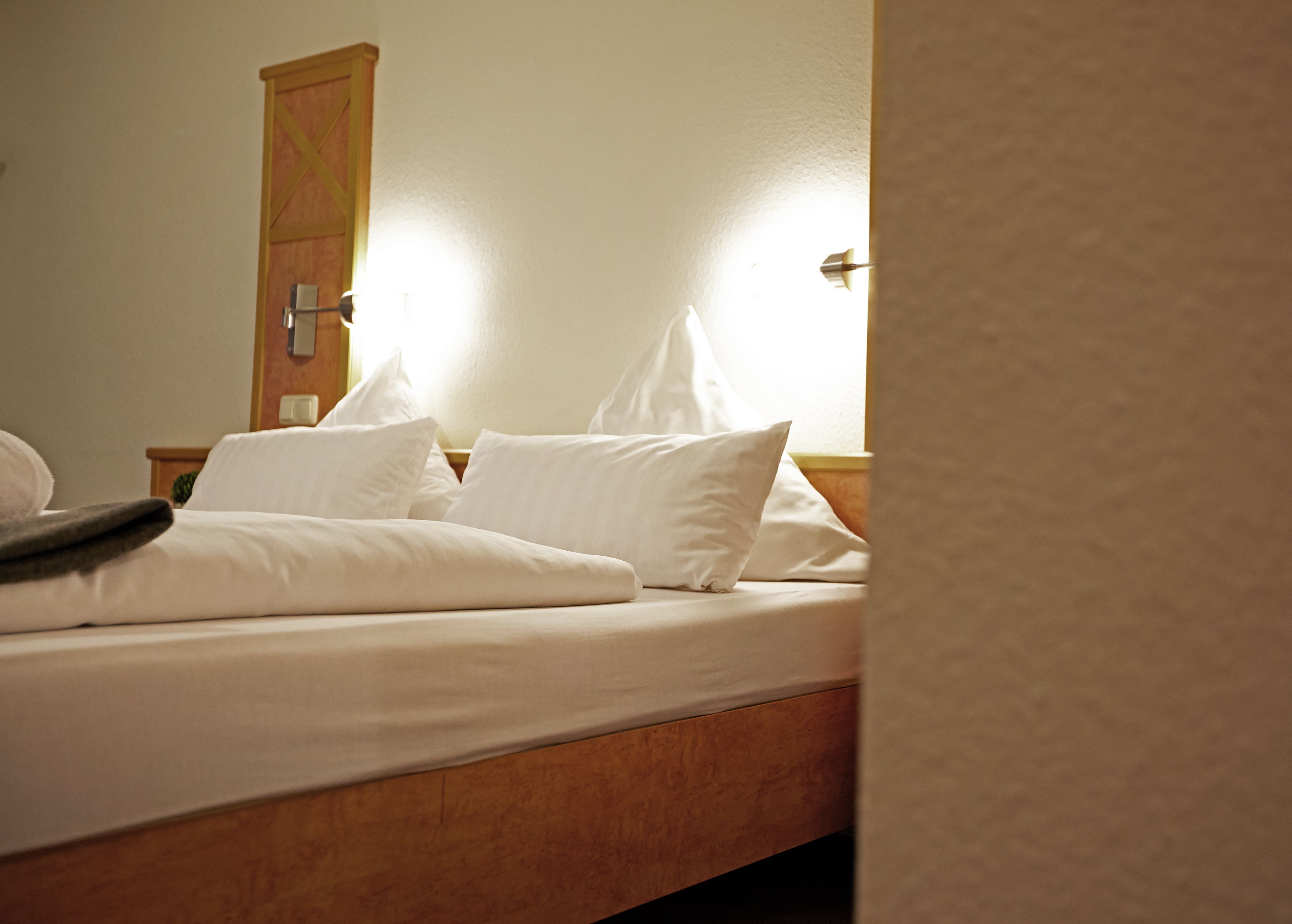 Un lit bien fait, avec du linge de lit blanc et des oreillers, une tête de lit en bois et un luminaire fixé au mur.