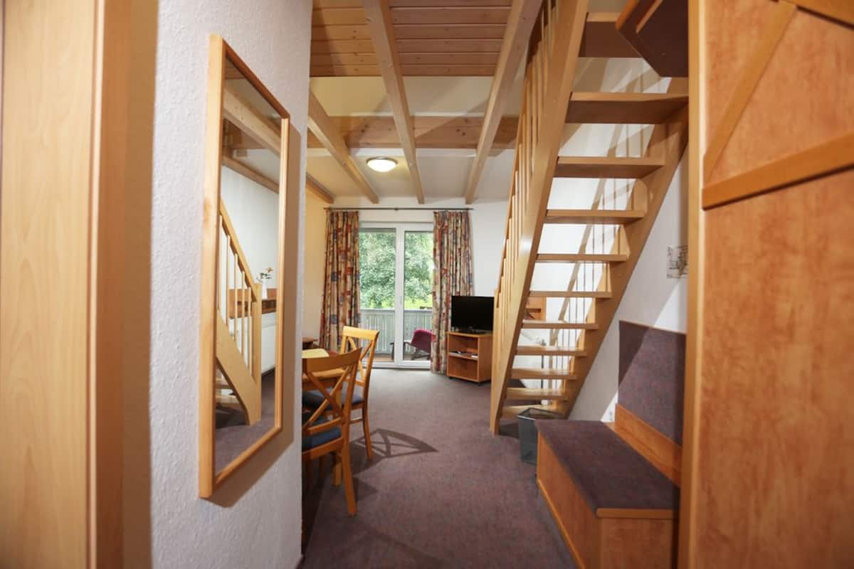 Un escalier en bois menant à un balcon, un coin repas avec des chaises et une table, et un téléviseur posé sur un meuble.